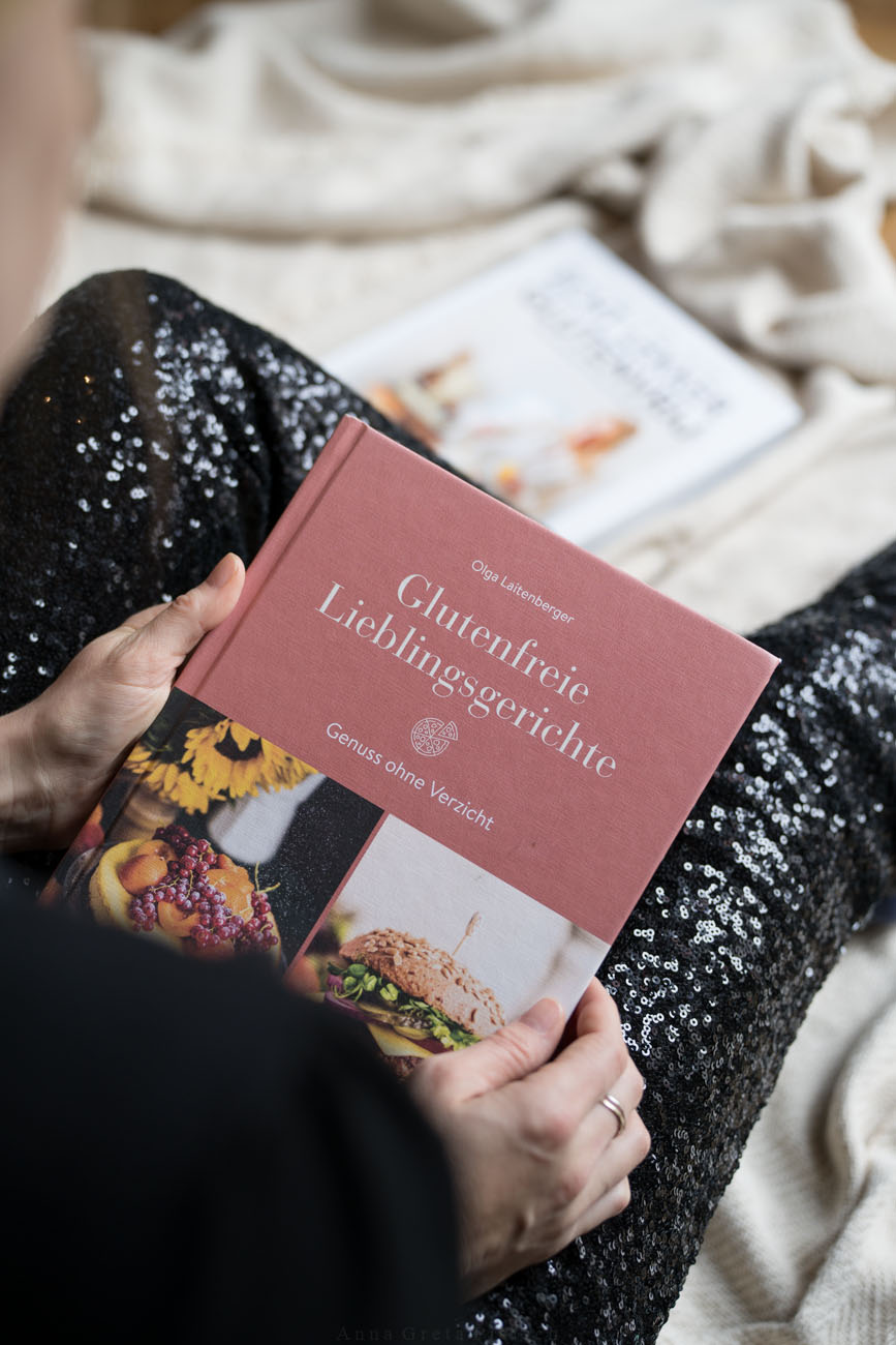 Eine Person sitzt in festlicher Kleidung (Pailetten-Hose) auf eier Decke auf dem Boden und hält ein glutenfreies Backbuch in den Händen.