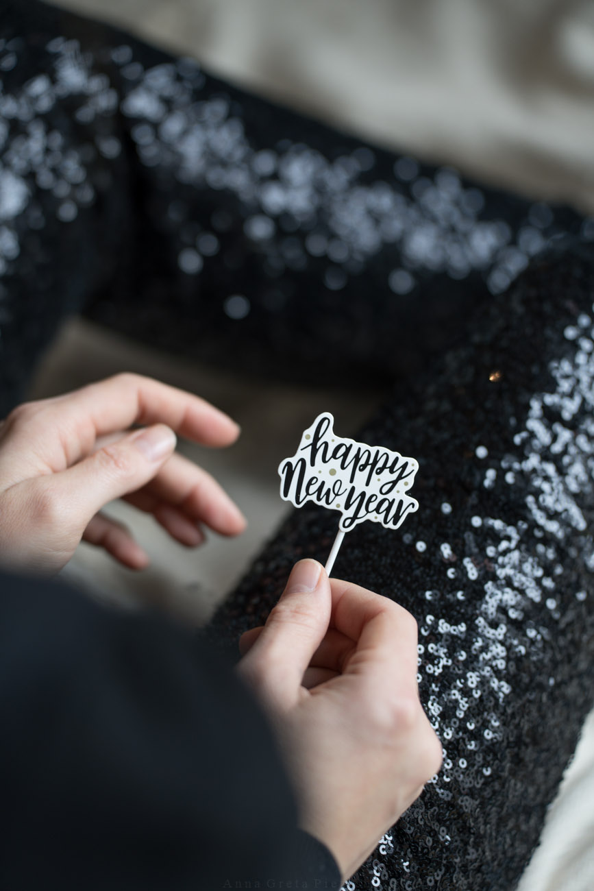 Eine Person sitzt in festlicher Kleidung (Pailetten-Hose) auf eier Decke auf dem Boden und hält einen Deko-Stecker mit "happy new year" in den Händen.