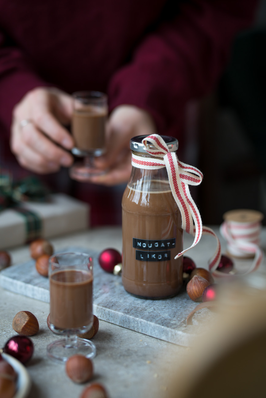 Ein kleines Fläschchen mit Nougat-Likör steht auf einem Marmorbrettchen. An der Flasche ist eine Schleife und der Tisch ist ein wenig weihnachtlich dekoriert. Im Hintergrung hält eine Person ein gefülltes Pinnchen ind en Händen.