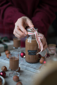 Ein kleines Fläschchen mit Nougat-Likör steht auf einem Marmorbrettchen. An der Flasche ist eine Schleife und der Tisch ist ein wenig weihnachtlich dekoriert.