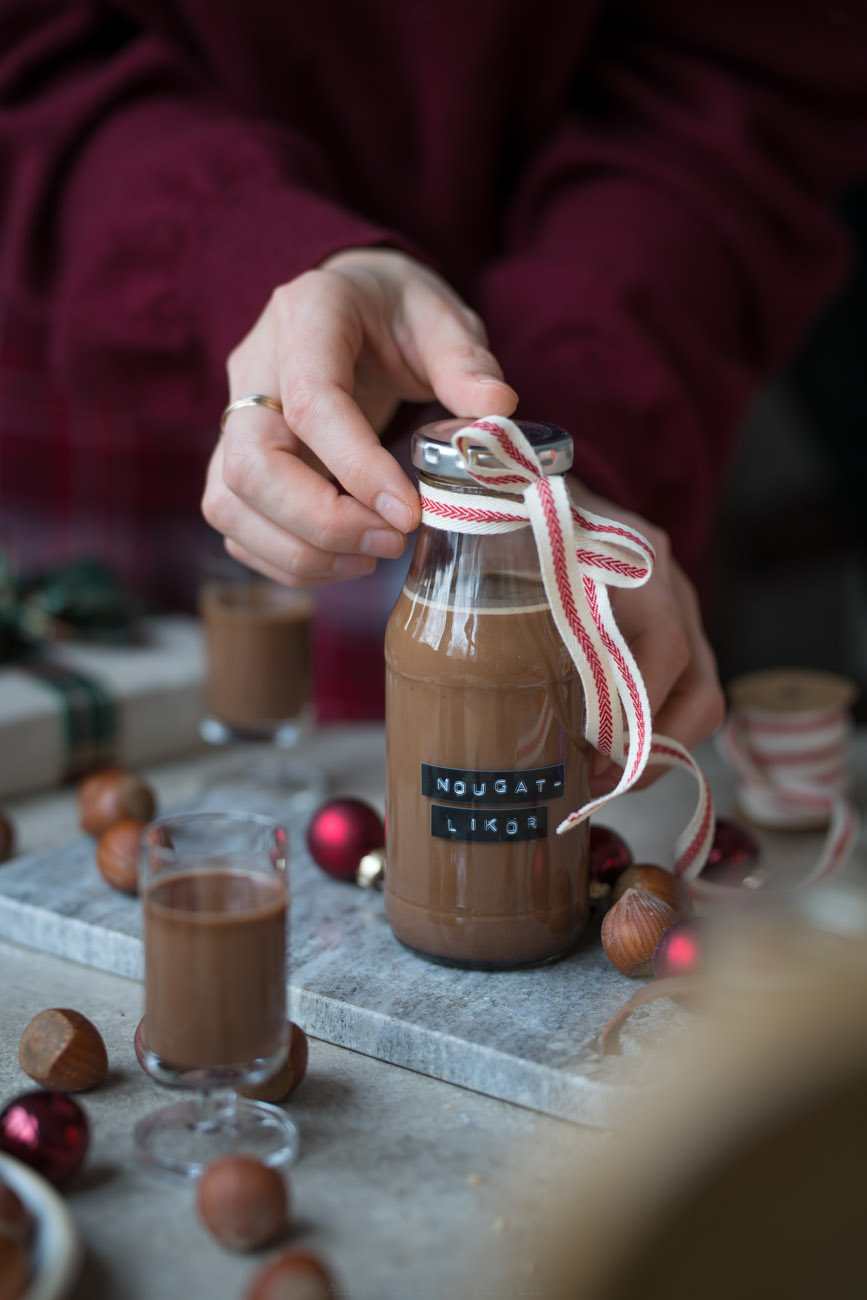 Ein kleines Fläschchen mit Nougat-Likör steht auf einem Marmorbrettchen. An der Flasche ist eine Schleie und der Tisch ist ein wenig weihnachtlich dekoriert.