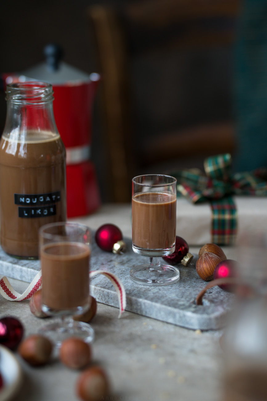 Ein Pinnchen mit Nougat-Likör auf einem Marmorbrettchen. Man sieht ein weiteres Pinnchen und eine kleine Flasche mit dem Likör. Der Tisch ist weihnachtlich geschmückt.