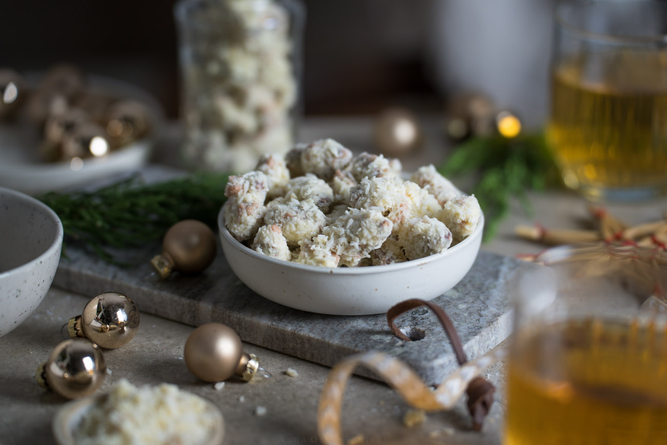 Ein winterlich gedeckter Tisch mit kleinen Weihnachtskugeln, etwas Tannengrün, Teegläsern. In der Mitte steht ein Schälchen, gefüllt mit gebrannten Mandeln, die in weißer Schokolade und Kokosstreuseln gewälzt wurden.