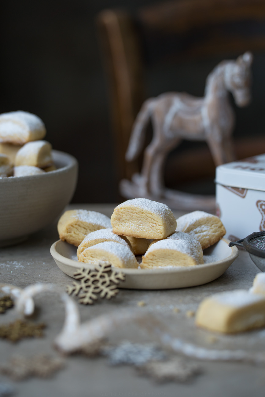 Ein schlicht weihnachtlich gedeckter Tisch. In der Mitte steht ein Teller mit Keksen, im Hintegrund ist eine Schale mit Keksen gefüllt zu sehen, Sonst steht ein Deko-Schaukelpferd, eine Keksdose auf dem Tisch.