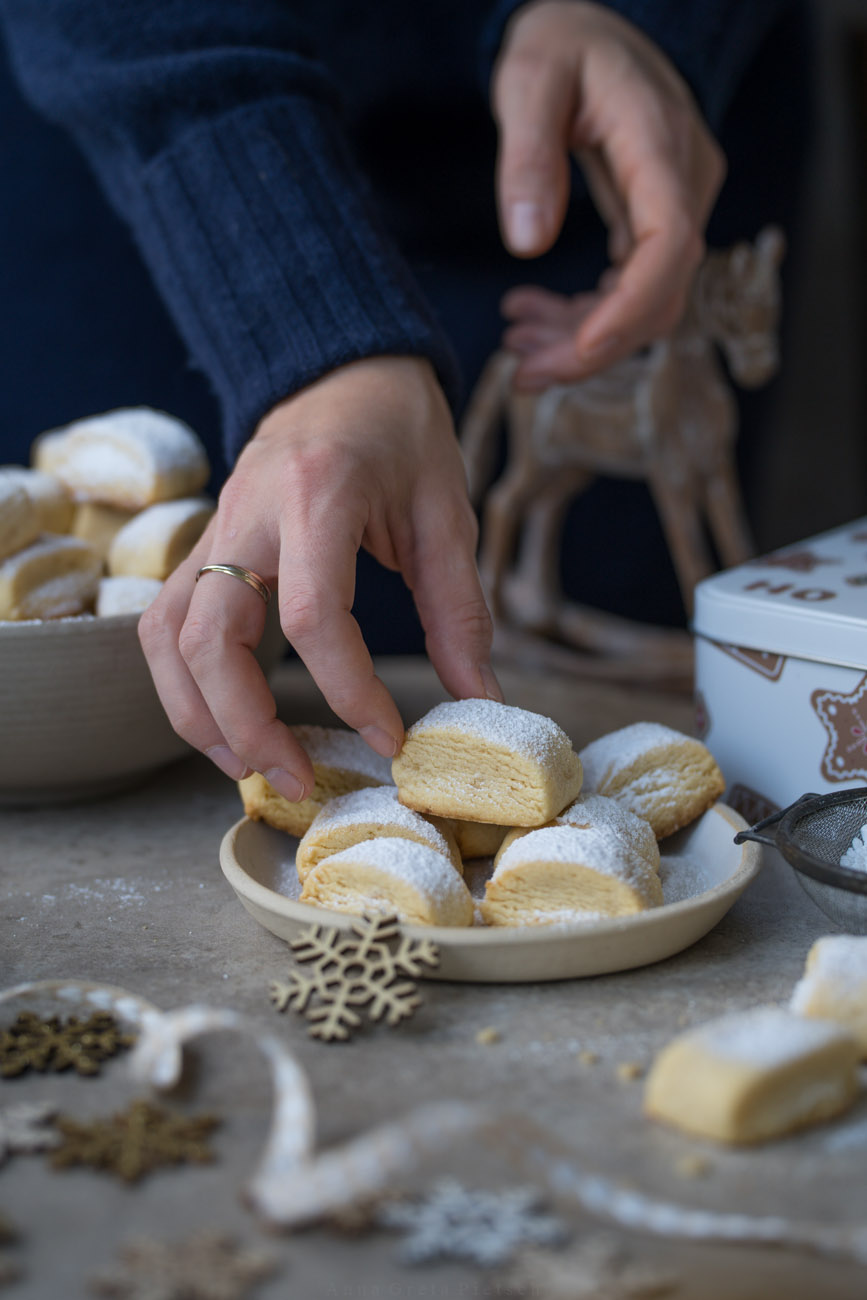 Ein schlicht weihnachtlich gedeckter Tisch. In der Mitte steht ein Teller mit Keksen, im Hintegrund ist eine Schale mit Keksen gefüllt zu sehen, Sonst steht ein Deko-Schaukelpferd, eine Keksdose auf dem Tisch. Eine Hand ist zu sehen, die sich ein Plätzchen wegnimmt. 