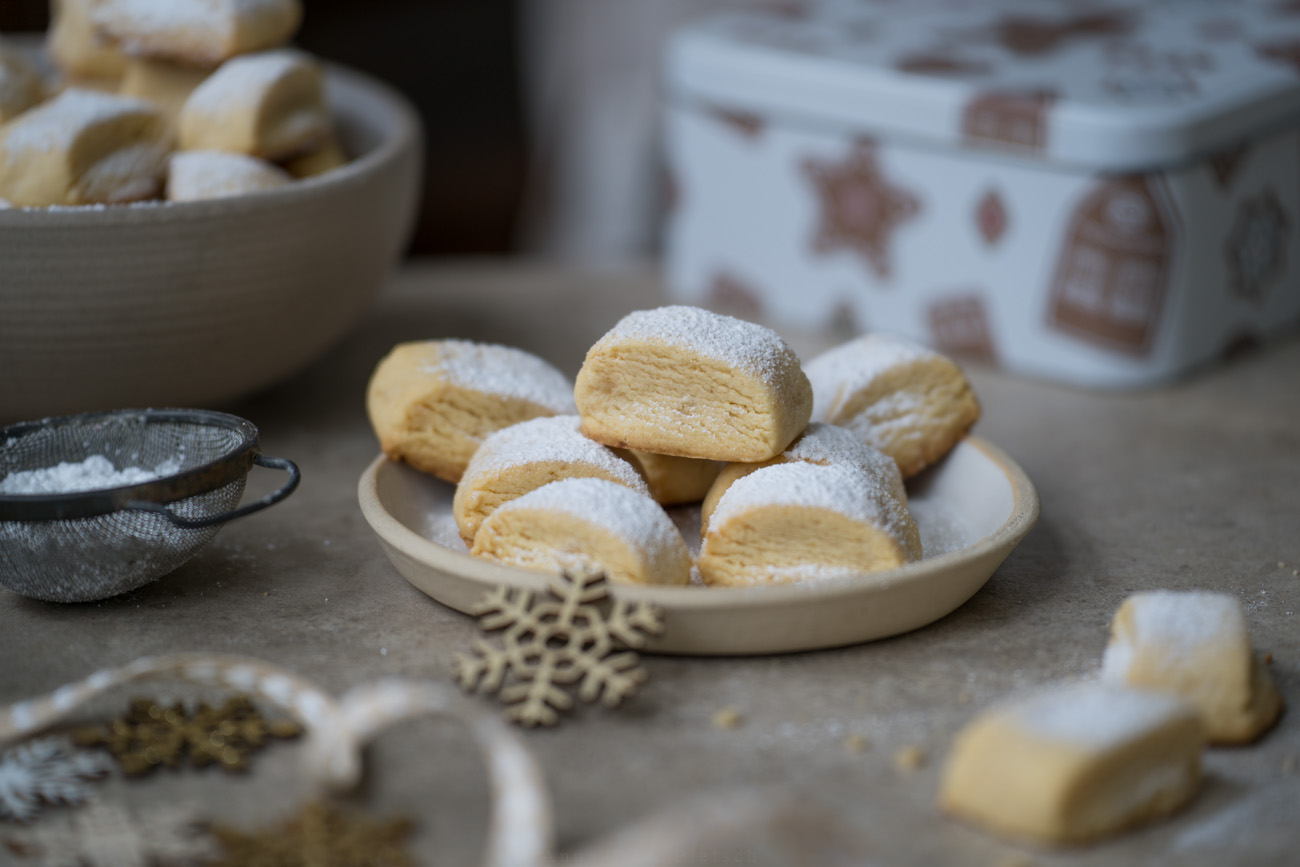 Ein schlicht weihnachtlich gedeckter Tisch. In der Mitte steht ein Teller mit Keksen, im Hintegrund ist eine Schale mit Keksen gefüllt zu sehen, Sonst steht eine Keksdose auf dem Tisch.