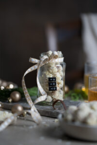 Ein winterlich gedeckter Tisch mit kleinen Weihnachtskugeln, etwas Tannengrün, Teegläsern. In der Mitte steht ein Schraubglas mit einer Schleife, gefüllt mit gebrannten Mandeln, die in weißer Schokolade und Kokosstreuseln gewälzt wurden.