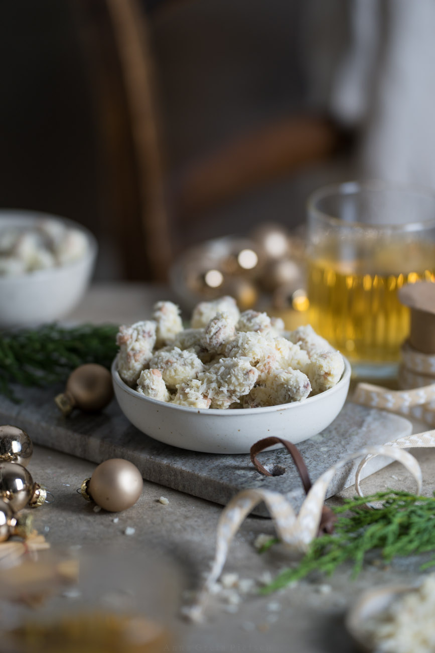 Ein winterlich gedeckter Tisch mit kleinen Weihnachtskugeln, etwas Tannengrün, Teegläsern. In der Mitte steht ein Schälchen, gefüllt mit gebrannten Mandeln, die in weißer Schokolade und Kokosstreuseln gewälzt wurden.