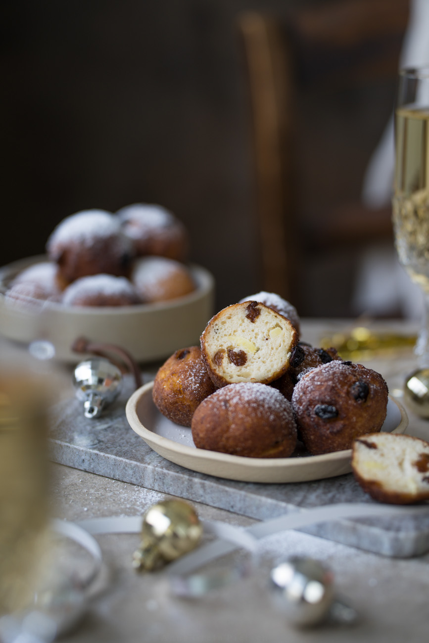 Ein Tisch mit einem Teller mit frittierten Teigbällchen in der Mitte. Man sieht auch eine entsprechend gefüllte Schale im Hintergrund stehen. Im Anschnitt sieht man vorne und hinten jeweils ein Sektglas.