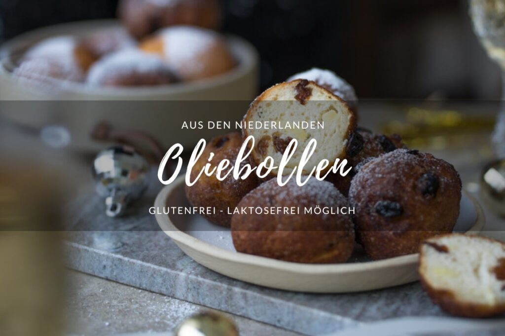 Rezept für Oliebollen. Ein Tisch mit einem Teller mit frittierten Teigbällchen in der Mitte. Man sieht auch eine entsprechend gefüllte Schale im Hintergrund stehen. Im Anschnitt sieht man vorne und hinten jeweils ein Sektglas.