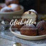 Rezept für Oliebollen. Ein Tisch mit einem Teller mit frittierten Teigbällchen in der Mitte. Man sieht auch eine entsprechend gefüllte Schale im Hintergrund stehen. Im Anschnitt sieht man vorne und hinten jeweils ein Sektglas.