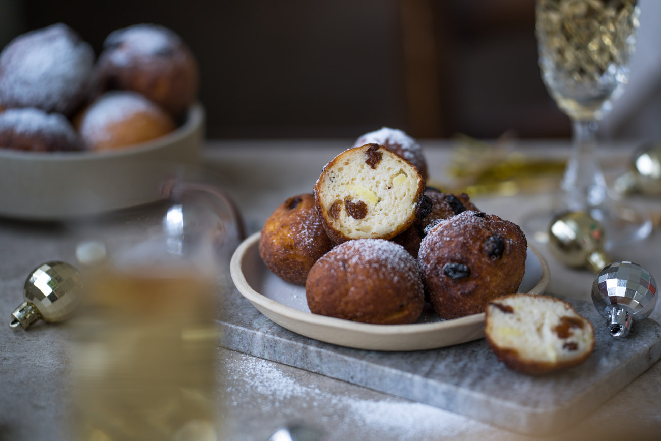 Ein Tisch mit einem Teller mit frittierten Teigbällchen in der Mitte. Man sieht auch eine entsprechend gefüllte Schale im Hintergrund stehen. Im Anschnitt sieht man vorne und hinten jeweils ein Sektglas.