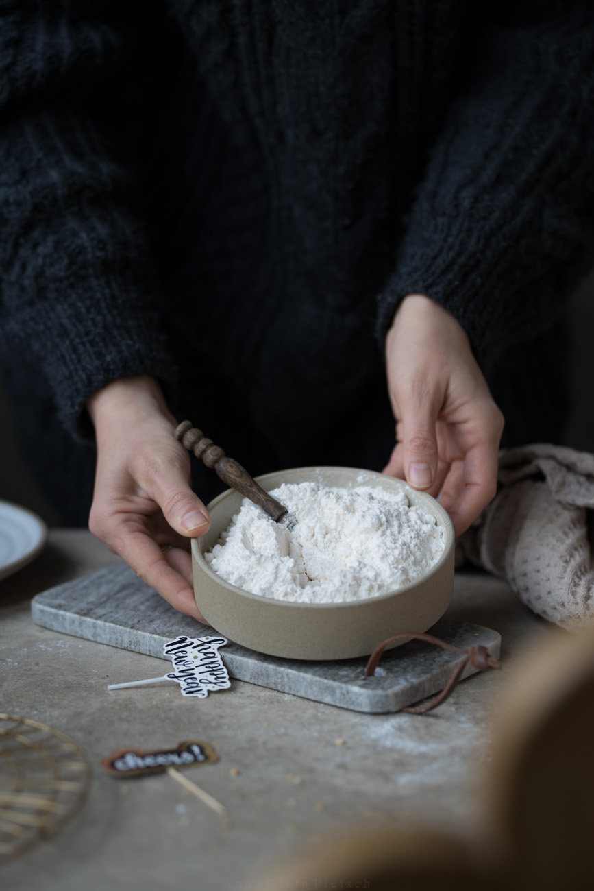 Eine Arbeitsfläche, in der Mitte wird Schale mit Mehl von zwei Händen gehalten.