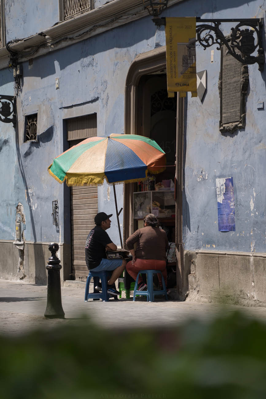 Zwei Personen sitzen auf Hockern vor einem Laden in Lima, Peru. Zwischen ihnen steht ein bunter Sonnenschirm. Die Hausfassade im Hintergrund ist schon in die Jahre gekommen und bröckelt ab.