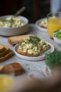 Ein gedeckter Frühstückstisch. In der Mitte steht ein kleiner Teller mit einer Brotscheibe, die mit selbstgemachtem veganem Eiersalat belegt ist.
