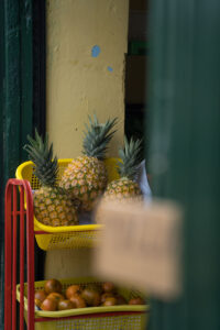 Ananas in einem Regal vor einem Geschäft. Die Hausfassade ist gelb, das Regal rot mit gelben Kisten.