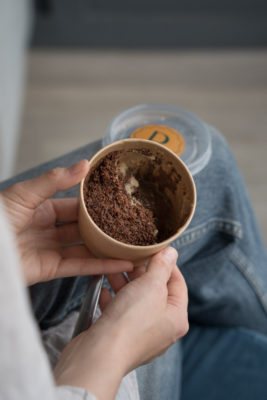 Eine Person hält eine Pappbowl mit einem bräunlichen Dessert in ihren Händen. Das Foto wurde über die Schulter aufgenommen.
