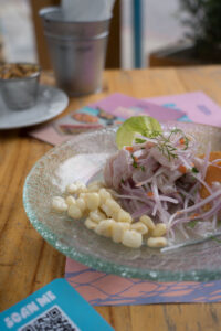 Ein Holztisch mit einem Glasteller. Darauf ist frisch zubereitetes Ceviche mit Maiskörnern als Beilage.