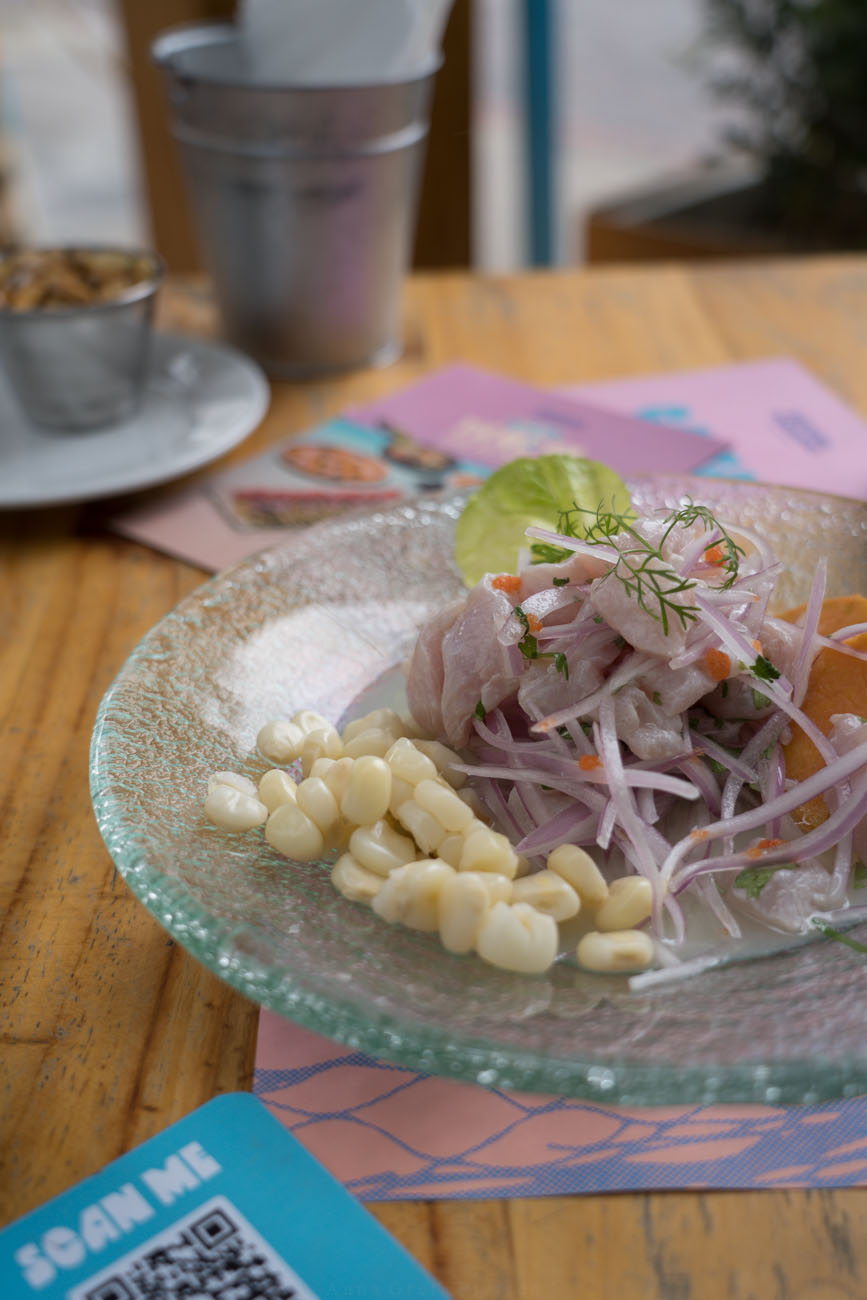 Ein Holztisch mit einem Glasteller. Darauf ist frisch zubereitetes Ceviche mit Maiskörnern als Beilage.