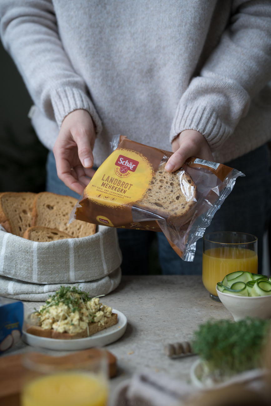 Ein gedeckter Frühstückstisch. Man sieht einen kleinen Teller mit einer Brotscheibe, die mit selbstgemachtem veganem Eiersalat belegt ist. Eine Person hält eine Brotpackung mit glutenfreiem Landbrot.