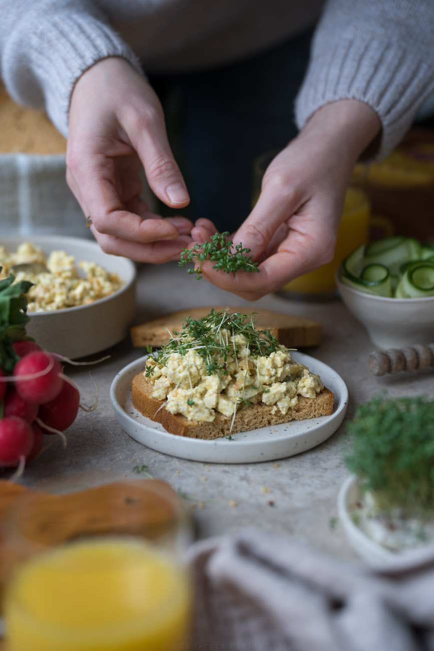 Ein gedeckter Frühstückstisch. In der Mitte steht ein kleiner Teller mit einer Brotscheibe, die mit selbstgemachtem veganem Eiersalat belegt ist. Zwei Hände bestreuen das Brot gerade noch mit frischer Kresse.