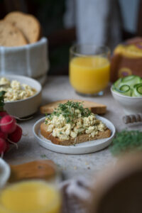 Ein gedeckter Frühstückstisch. In der Mitte steht ein kleiner Teller mit einer Brotscheibe, die mit selbstgemachtem veganem Eiersalat belegt ist.