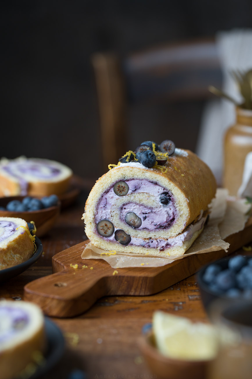 Man sieht einen Holztisch, daraug steht in der Mitte eine Biskuitrolle mit einer lilafarbenen Füllung aus Blaubeeren. Man erkennt in der Füllung auch frische Blaubeeren im Anschnitt.
