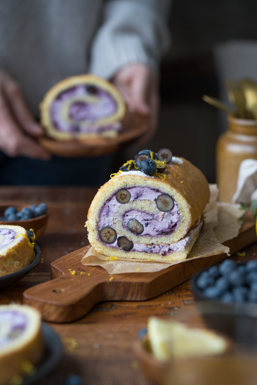 Man sieht einen Holztisch, daraug steht in der Mitte eine Biskuitrolle mit einer lilafarbenen Füllung aus Blaubeeren. Man erkennt in der Füllung auch frische Blaubeeren im Anschnitt. Im Hintergrund hält eine Person ein Stück des Kuchens auf einem Teller in den Händen.