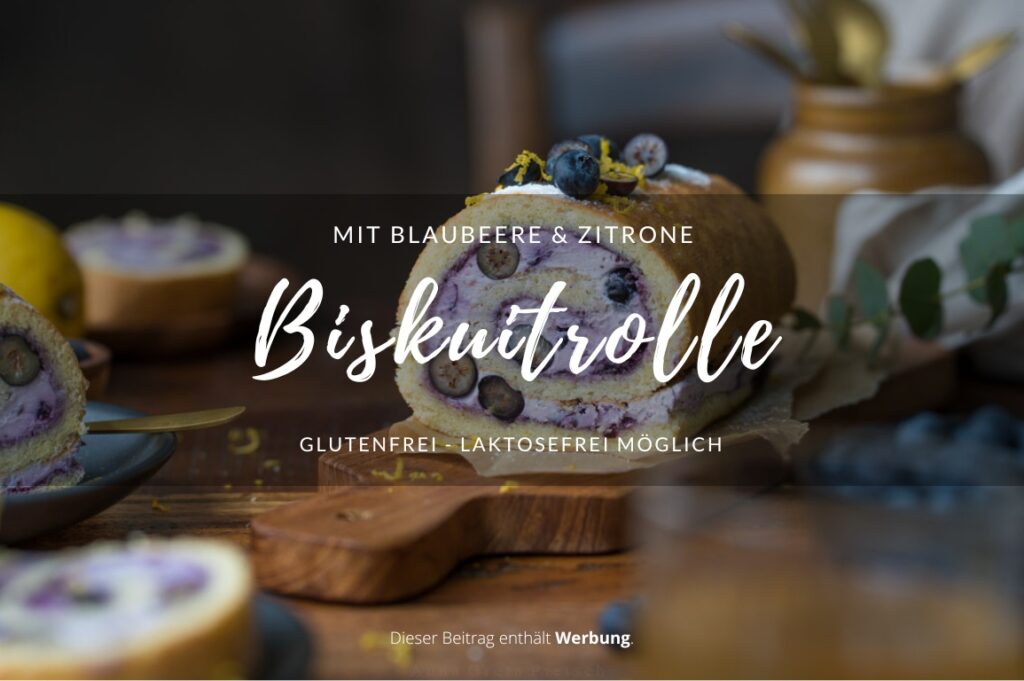 Man sieht einen Holztisch, daraug steht in der Mitte eine Biskuitrolle mit einer lilafarbenen Füllung aus Blaubeeren. Man erkennt in der Füllung auch frische Blaubeeren im Anschnitt.