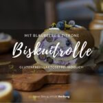 Man sieht einen Holztisch, daraug steht in der Mitte eine Biskuitrolle mit einer lilafarbenen Füllung aus Blaubeeren. Man erkennt in der Füllung auch frische Blaubeeren im Anschnitt.