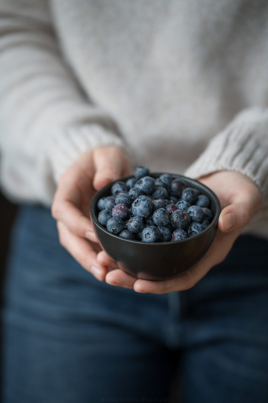 Eine Person hält eine schwarze Schale in beiden Händen. Die Schale ist mit Blaubeeren gefüllt. Auf den Beeren sind noch Wassertropfen zu erkennen.