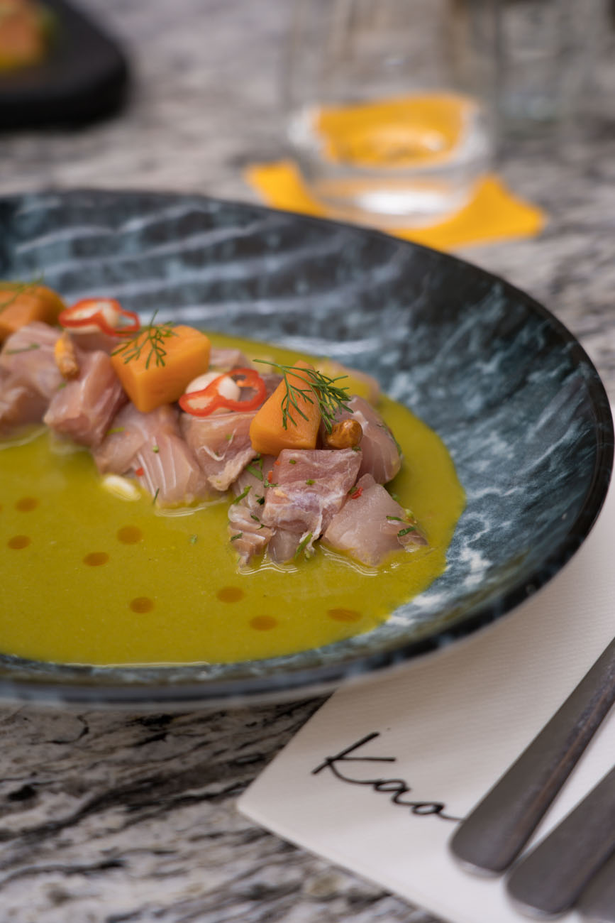 Ein Tisch aus Marmor in einem Restaurant. Darauf sieht man im Hintergrund unscharf ein Wasserglas und im Vordergrund einen tiefen Teller in dunklen Farben mit Ceviche gefüllt.