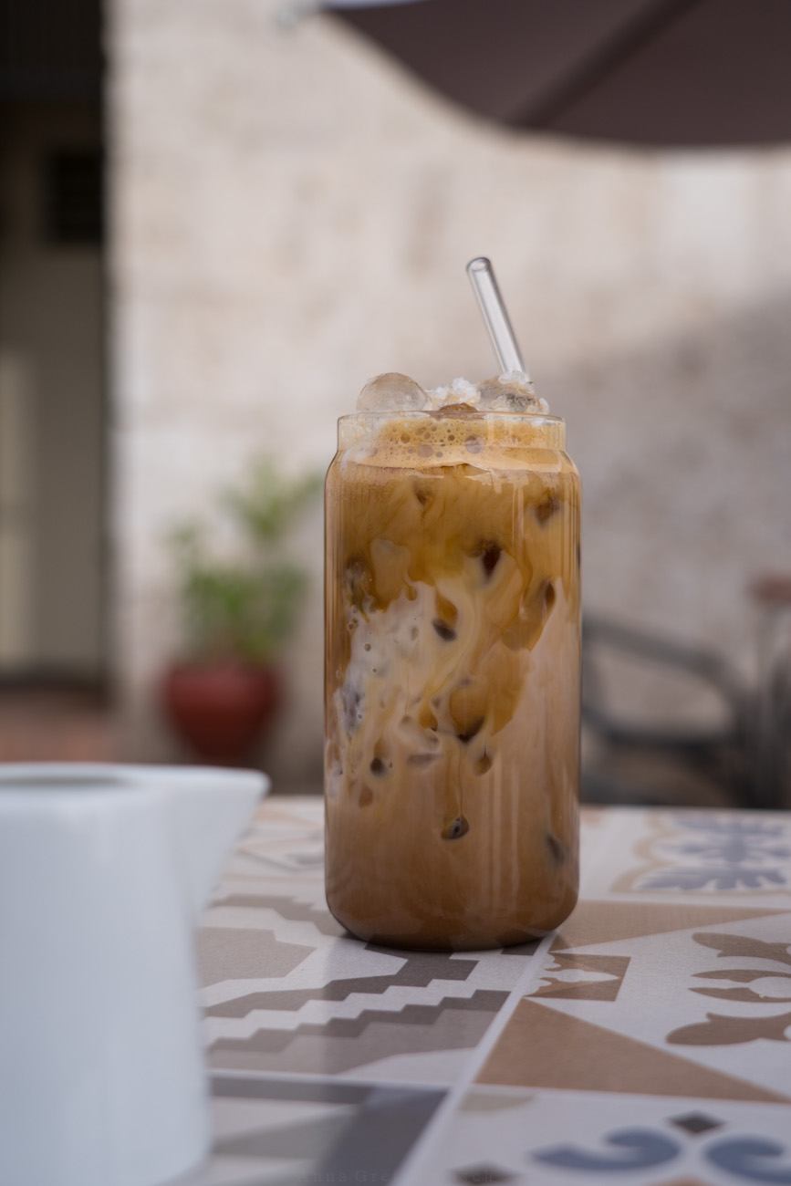 Man sieht ein hohes Glas auf einem Tisch in einem Café. Das Glas ist mit Eiskaffee geüllt. Darin steckt ein Glasstrohhalm.