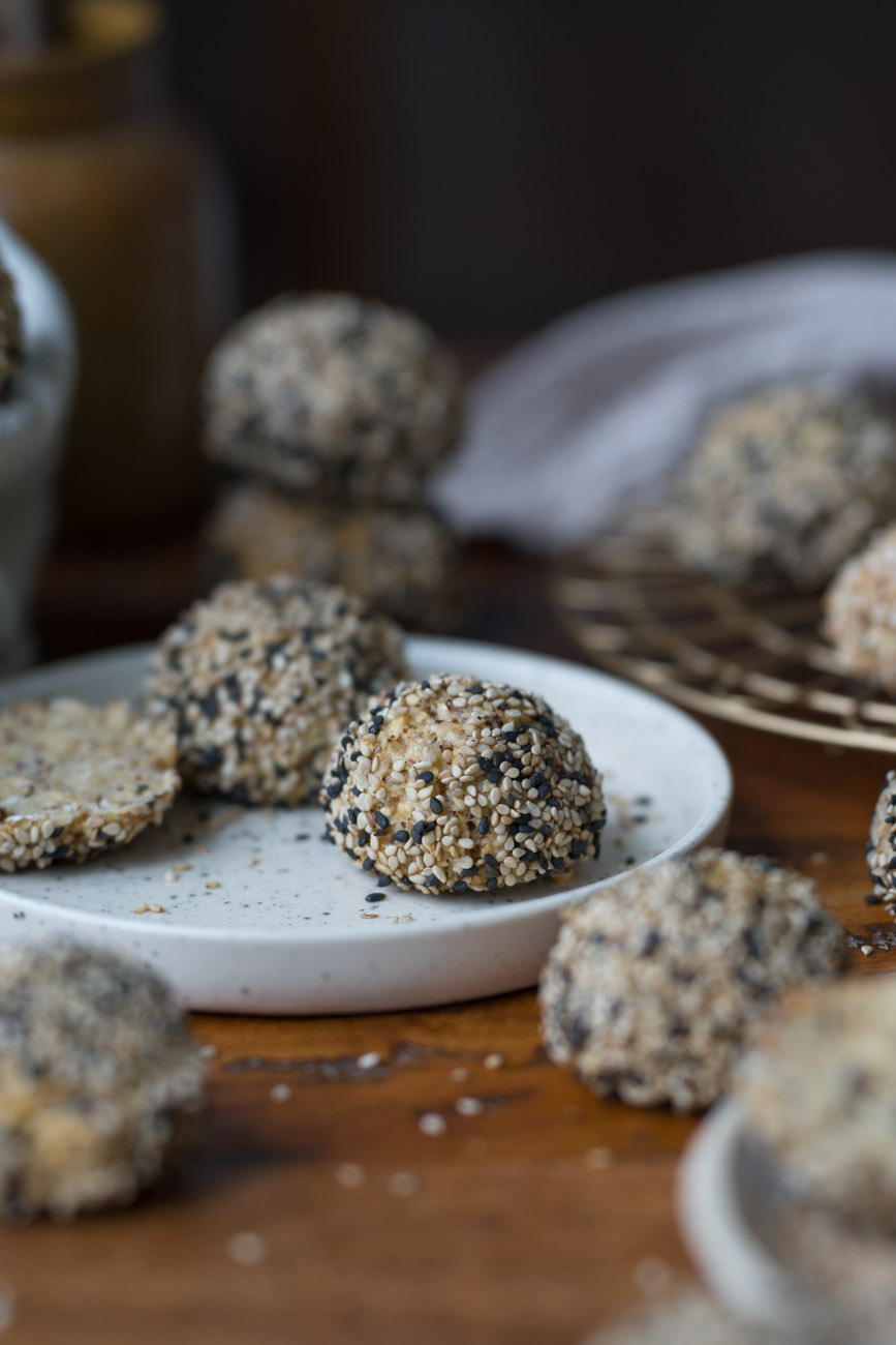 Eine Frühstücksszene. Ein kleiner Teller steht auf einer Holzplatte. Darauf liegt ein Mini-Brötchen mit hellen und dunklen Sesam-Samen.