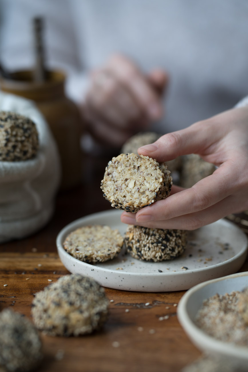 Eine Frühstücksszene. Ein kleiner Teller steht auf einer Holzplatte. Darauf liegt ein Mini-Brötchen mit hellen und dunklen Sesam-Samen. Eine Hand hält ein aufgeschnittenes Mini-Brötchen ins Bild.