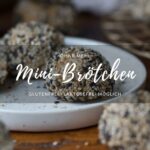 Eine Frühstücksszene. Ein kleiner Teller steht auf einer Holzplatte. Darauf liegt ein Mini-Brötchen mit hellen und dunklen Sesam-Samen. Ein Titelbild für ein Rezept.