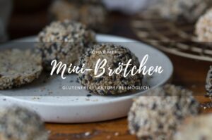 Eine Frühstücksszene. Ein kleiner Teller steht auf einer Holzplatte. Darauf liegt ein Mini-Brötchen mit hellen und dunklen Sesam-Samen. Ein Titelbild für ein Rezept.