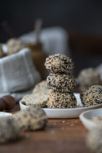 Eine Frühstücksszene. Ein kleiner Teller steht auf einer Holzplatte. Darauf sind drei Mini-Brötchen mit hellen und dunklen Sesam-Samen gestapelt.