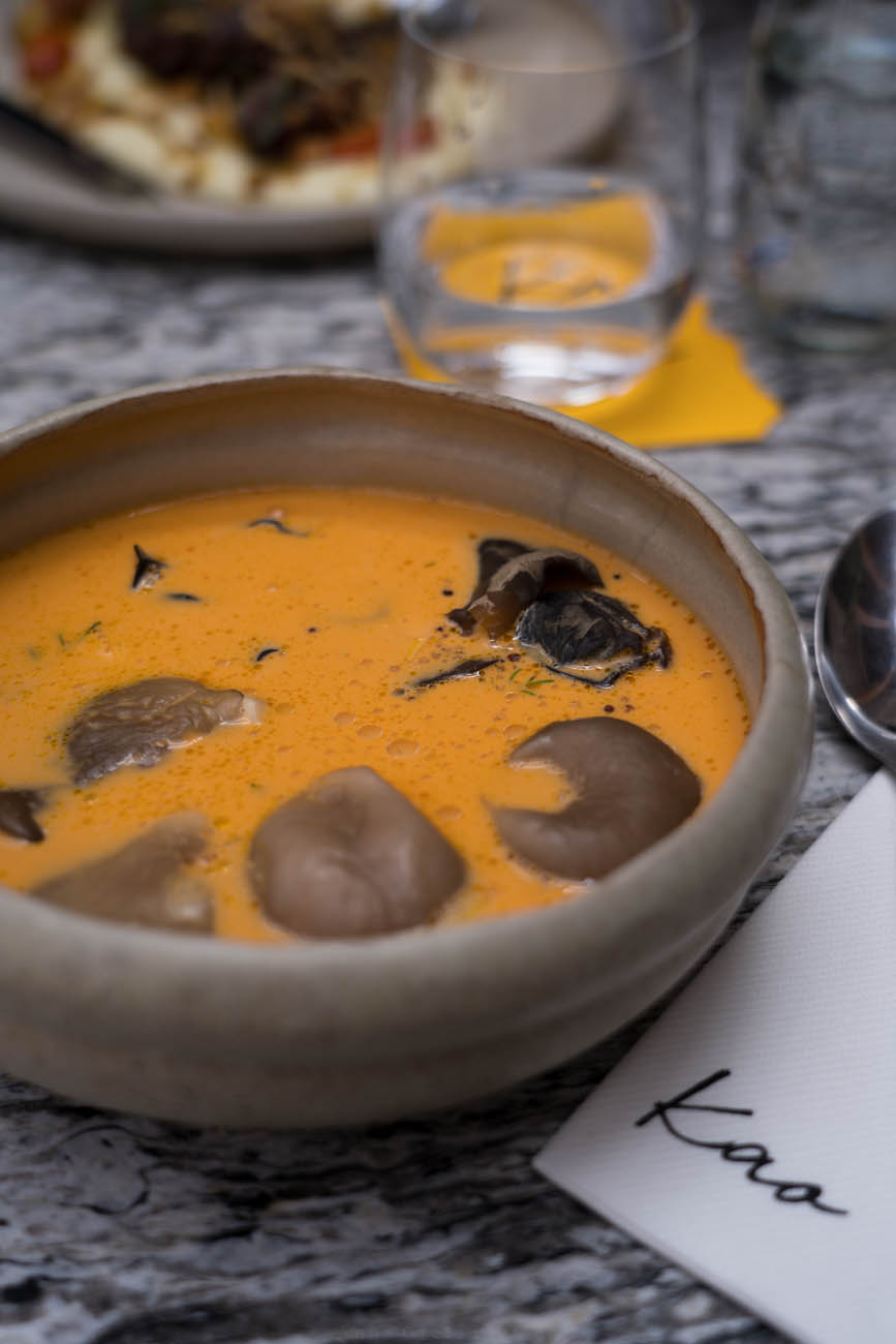 Ein Tisch aus Marmor in einem Restaurant. Darauf sieht man im Hintergrund unscharf ein Wasserglas und im Vordergrund eine beige Suppenschale mit einer orangefarbenen Suppe mit Pilzen darin.