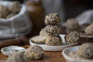 Eine Frühstücksszene. Ein kleiner Teller steht auf einer Holzplatte. Darauf sind drei Mini-Brötchen mit hellen und dunklen Sesam-Samen gestapelt.