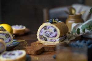 Man sieht einen Holztisch, darauf steht in der Mitte eine Biskuitrolle mit einer lilafarbenen Füllung aus Blaubeeren. Man erkennt in der Füllung auch frische Blaubeeren im Anschnitt.