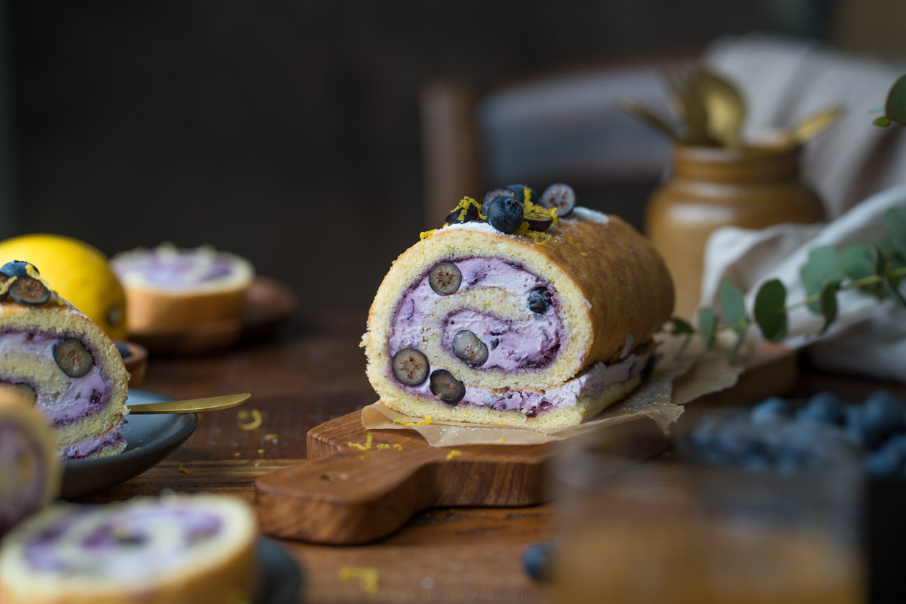 Man sieht einen Holztisch, daraug steht in der Mitte eine Biskuitrolle mit einer lilafarbenen Füllung aus Blaubeeren. Man erkennt in der Füllung auch frische Blaubeeren im Anschnitt.