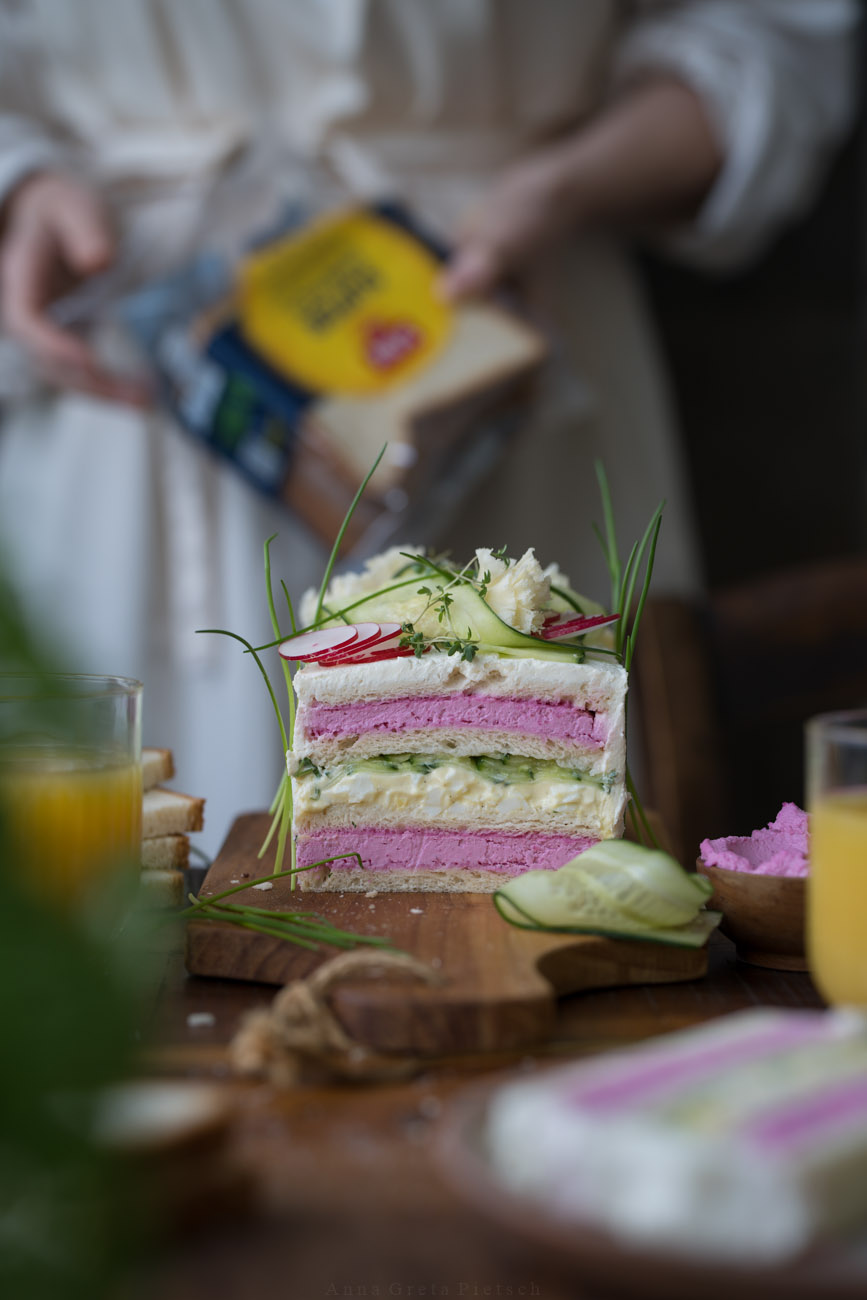 Man sieht einen Holztisch. Er ist für ein Frühstück gedeckt. Man sieht Orangensaftgläser und in der Mitte eine Sandwichtorte mit verschiedenen Schichten. Im Hintergrund hält eine Person eine Brotverpackung in den Händen. 