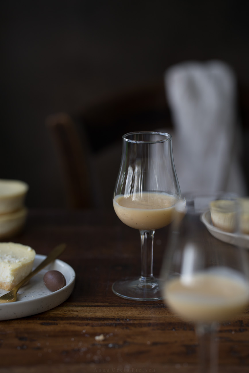 Ein Glas Eierliköt steht auf einer dunklen Holztischplatte. Man sieht Teller mit Kuchen im Hintergrund.