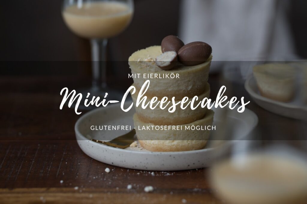 Drei gestapelte Mini-Cheesecake (in Muffingröße) stehen auf einem kleinen Teller auf einem Holztisch. Im Vorder- und Hintergrund erkennt man jeweils ein Glas Eierlikör.
