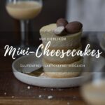 Drei gestapelte Mini-Cheesecake (in Muffingröße) stehen auf einem kleinen Teller auf einem Holztisch. Im Vorder- und Hintergrund erkennt man jeweils ein Glas Eierlikör.