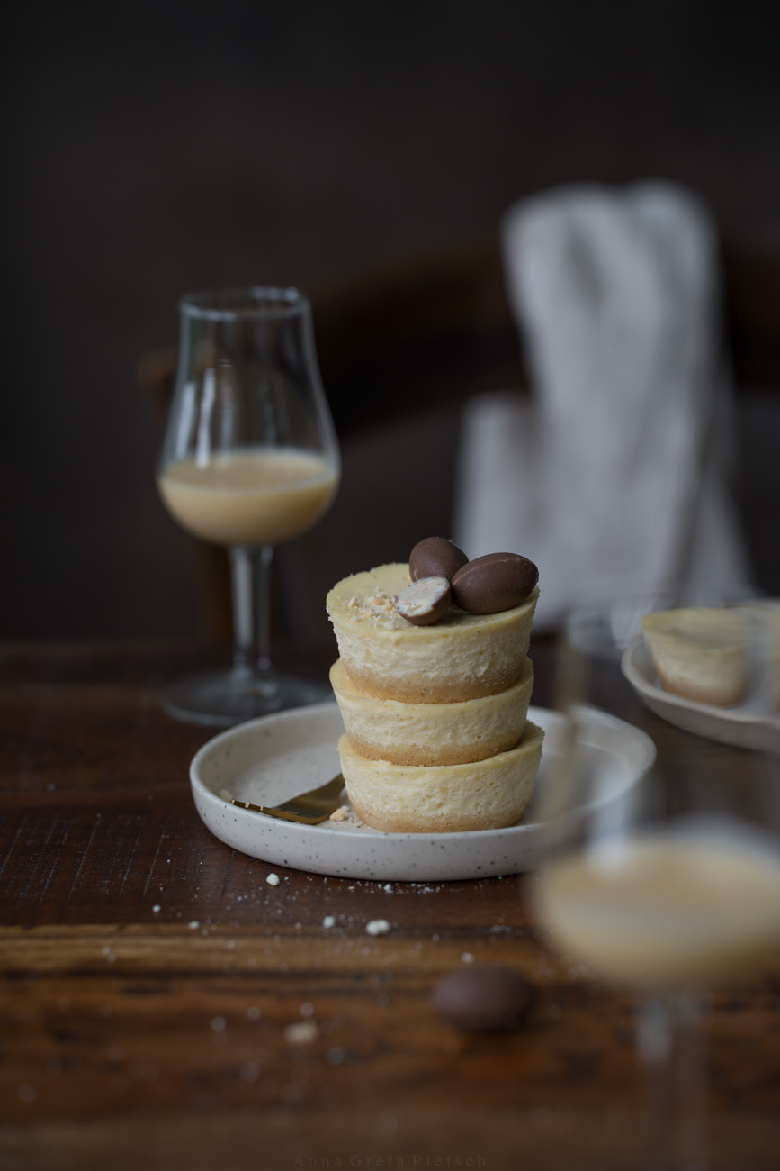Drei gestapelte Mini-Cheesecake (in Muffingröße) stehen auf einem kleinen Teller auf einem Holztisch. Im Vorder- und Hintergrund erkennt man jeweils ein Glas Eierlikör.