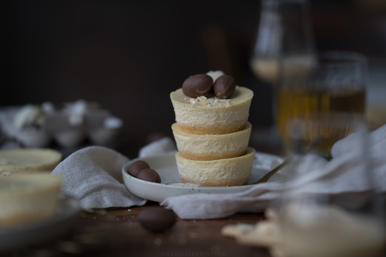 Drei gestapelte Mini-Cheesecake (in Muffingröße) stehen auf einem kleinen Teller auf einem Holztisch. Im Vorder- und Hintergrund erkennt man jeweils ein Glas Eierlikör.