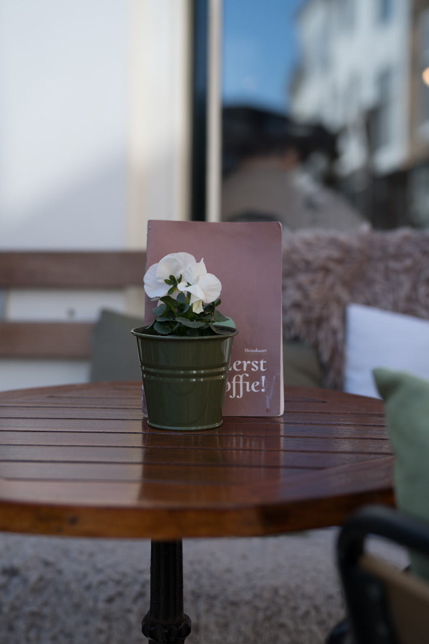 Ein Tisch mit einer Holzplatte auf der Außenterrasse eines Cafés. Auf dem Tisch steht eine Pflanze in einem kleinen grünen Blechtopf.