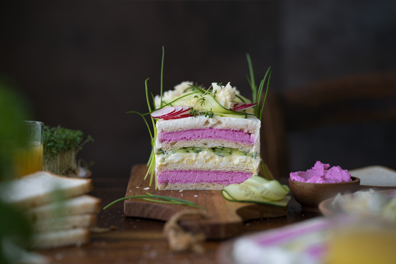 Man sieht einen Holztisch. Er ist für ein Frühstück gedeckt. Man sieht Orangensaftgläser und in der Mitte eine Sandwichtorte mit verschiedenen Schichten.